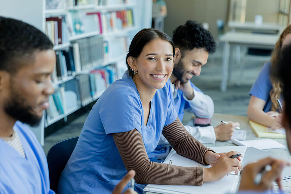 Three nursing students smile as they take notes in a classroom.